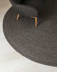 The Armadillo&Co Braid Rug - Charcoal, a round wool-blend rug, rests on light wood flooring beneath a dark gray chair with light wooden legs.