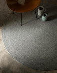 The Armadillo&Co Braid Rug - Heron, a round handmade gray wool-blend rug, rests on a concrete floor with a wooden chair leg, white mug, and blue ceramic pitcher arranged at its edge.