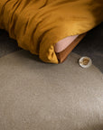 A bed with a mustard yellow blanket, the Armadillo&Co Braid Rug - Mushroom (handmade from wool-blend yarn) on a speckled floor, and a white cup on a saucer placed next to the bed.