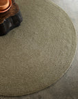 The Braid Rug - Oregano by Armadillo&Co, a round handmade piece in beige, rests on a speckled concrete floor. Part of a dark wooden coffee table with a terracotta dish appears in the upper left corner.