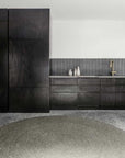 Modern kitchen with dark wood cabinets, matte gray tile backsplash, gold faucet, decorative vases on the counter, and the Armadillo&Co Braid Rug - Pumice on a speckled floor. The design is minimal and clean.