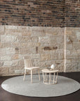 A light wooden chair and small round table with a teapot and cup sit on the Armadillo&Co Braid Rug in Pumice in front of a stone and brick wall on a dark wooden floor.