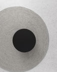 The Armadillo&Co Braid Rug - Pumice, a textured light gray wool-blend, lies on a speckled white surface with a black circular object at its center, viewed from above.