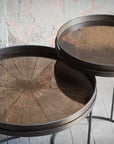 Two Ethnicraft Bronze Mirror Tray - Round / Small tables with textured bronze surfaces and slim black legs stand together on an old wood floor by a distressed plaster wall, radiating refined European style.