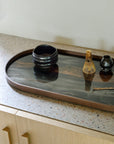 Part of Ethnicraft’s Tray Collection, the Bronze Organic Glass Tray (Oblong/Medium) features a dark, reflective surface and sits on a terrazzo countertop above light wood cabinets. It holds a black bowl, bamboo whisk, jug, and metal spoon.