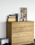 An Ethnicraft Burger Dresser, crafted from solid European oak, stands against a white wall with stacked books, decor, and a grid ornament on top. Magazines and black shoes rest on the floor beside it.