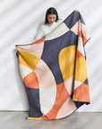 A woman in a white sweater holds up the Sunset Quilt by FAIRE/ANCHAL, featuring hand-stitched abstract geometric patterns in navy, yellow, peach, cream, and orange, standing against a plain white wall.