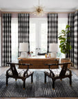 A modern home office features a wooden desk, checkered chairs, matching lamps, a potted plant, and floor-to-ceiling plaid curtains. The Loloi Cadence Rug in Charcoal adds warmth beneath a striking gold starburst chandelier.