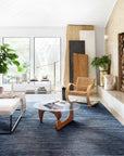A modern living room with large windows, indoor plants, a woven chair, and a glass coffee table. Decorative wood panels lean against the wall, while a fireplace sits on the right. The Loloi Cadence Rug - Navy covers the floor.