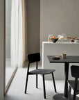 A minimalist dining area showcases the Ethnicraft Casale Dining Chair - Black and a European oak table with a cup and small plate. In the background, jars of oranges, pastries under glass, and a bowl are softly illuminated by natural light.
