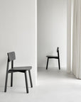 Two Casale Dining Chairs in black by Ethnicraft are positioned in a bright, white room with smooth floors and neutral walls; one is closer to the foreground, while the other rests near a doorway, ready to complement any dining table.