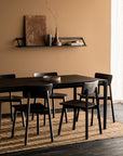 A minimalist dining room features six Ethnicraft Casale Dining Chairs in black with a matching table on a woven rug, beige walls, a floating shelf with ceramics and a book, and textured art, softly lit by natural light from the right.