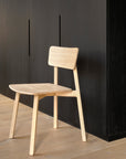 The Casale Dining Chair - Oak by Ethnicraft, with its simple lines and light European oak finish, sits on a pale wood floor before a tall dark cabinet, showcasing Ethnicraft’s minimalist style and classic European refinement.