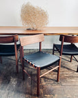 A wooden dining table with three SOBU Cesco Dining Chairs in walnut, each with black top grain leather seats, sits on a rustic wooden floor. A dried tumbleweed centerpiece decorates the tabletop.