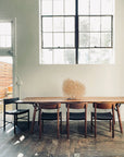 Minimalist dining area with a long Eastern black walnut table, four wooden chairs, one SOBU Cesco Dining Chair - Walnut in top grain leather, and a twig centerpiece. Large windows brighten the space, highlighting wood floors and white walls.