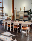 A sunlit room features a long wooden table and eight SOBU Cesco Dining Chairs in White Ash, decorative wooden objects on the table, rugs displayed on ladders, a plant, shelves with items, and wooden floors.