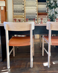 Two SOBU Cesco Dining Chairs in White Ash with top grain leather seats sit at a table in a showroom, with fabric samples displayed on the wall behind them and a potted plant on the right.