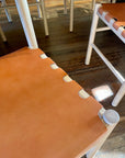 A close-up of SOBU Cesco Dining Chairs in White Ash with orange top grain leather seats, visible notches at the leather attachment points, set on a rustic wooden floor bathed in sunlight.