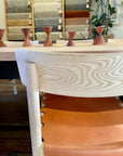 A close-up of the SOBU Cesco Dining Chair – White Ash with a tan top grain leather seat at a wooden table. In the background, five terracotta candleholders, fabric samples, and a plant are visible on the wall.