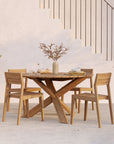 An Ethnicraft Circle Outdoor Dining Table in natural teak with four matching chairs sits by a staircase, styled with plates, cups, books, and a vase of dried branches. Sunlight casts soft shadows on this FSC-certified teak set.