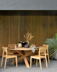 A modern outdoor dining area features Ethnicrafts Circle Outdoor Dining Table in natural teak with four matching chairs, set with dishes and glasses. Dried plants and a palm provide natural accents against a vertical wooden slat wall.