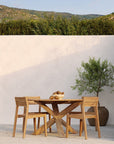 The Ethnicraft Circle Outdoor Dining Table in natural teak, paired with three matching chairs and a bowl of fruit, sits on a patio. A potted olive tree is nearby, with green shrubs and distant hills in the background.