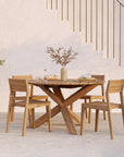 The Ethnicraft Circle Outdoor Dining Table in natural teak, FSC certified, sits by a staircase with four matching chairs. The table displays a vase of dried branches, dishes, bread, and drinks. An olive tree in a pot stands against a light wall nearby.