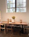 A wooden dining table with six assorted chairs, including the SOBU Copenhagen Dining Chair - White Ash, sits by a large window. The solid wood table and neutral pottery enhance the sunlit, minimalist decor.