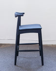 The SOBU Copenhagen Counter Stool - Black Ash features a curved backrest and top-grain black leather seat, set on a round textured gray rug against a plain white wall.