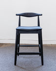 The SOBU Copenhagen Counter Stool - Black Ash features a modern curved backrest and a dark padded top-grain black leather seat, shown on a gray textured rug against a plain white wall.