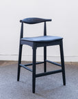 The SOBU Copenhagen Counter Stool - Black Ash features a curved backrest and square seat, standing on a round gray rug against a plain white wall and dark wood floor.