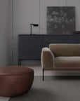 A minimalist living room with a beige velvet sofa, the Wooden Stories Fable Ottoman in leather as extra seating, a dark sideboard, small lamp, bowl, blurred artwork against light gray walls, and a large window.