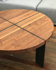 The SOBU Dial Coffee Table - Walnut features a round eastern walnut top and two black metal legs in a cross pattern, shown on a wooden floor next to a gray sofa.