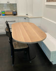 A SOBU Disco Racetrack Dining Table in white oak with a patina steel base sits with black metal chairs and a built-in white bench against white cabinets and dark floors, while a colorful toy and digital clock are on the counter.
