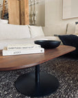 A modern living room features the SOBU Disco Racetrack Coffee Table in black walnut with a patina steel base, topped with two books and a black bowl. A light sofa with cushions sits behind, and a textured rug lies beneath the table.