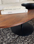 Two books, “Everything I Want to Eat” and “AD at 100,” are stacked on the SOBU Disco Racetrack Coffee Table in black walnut, which stands on a woven rug with a black bowl nearby and a beige sofa in the background.