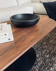 A modern living room featuring the SOBU Disco Racetrack Coffee Table in black walnut with a patina steel base, matte black bowl, and stacked books on a textured knit rug before a light sofa. Custom sizes and finishes available.