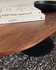 A close-up of the SOBU Disco Racetrack Coffee Table in Black Walnut with a patina steel base, topped with two stacked books—Everything I Want to Eat and AD at 100—and a black bowl on a textured woven rug.