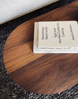 Two books sit atop SOBU’s Disco Racetrack Coffee Table in Black Walnut, beside a gray textured rug. The table features a solid walnut top and patina steel base. Custom sizes and finishes are available.