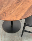The Disco Round Dining Table - Black Walnut by SOBU features striking wood grain and a black metal base, paired here with a black wooden chair on concrete. Custom sizes and finishes are available to suit your space.