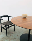 A black wooden chair sits beside the SOBU Disco Round Dining Table in Eastern black walnut with a black pedestal base. Three cream bowls are stacked on the table, and the minimalist setting showcases signature SOBU design.