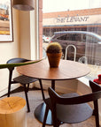 A SOBU Disco Round Dining Table in black walnut with a potted cactus sits by a café window. Two black chairs are at the table, and “The Levant” restaurant sign is visible outside.