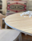A close-up of the Disco Round Dining Table in White Ash by SOBU, featuring a soft, rounded edge. A white decorative object sits on top, with a blurred patterned cushion and wooden chair in the background.