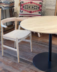 A light wood chair in American ash with a curved backrest sits next to the SOBU Disco Round Dining Table in white ash with a black metal base. Behind them are textiles, including a geometric cushion in red and black.
