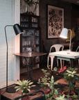 A cozy indoor space with potted plants, two tables featuring Seed Lightings Dodo Floor Lamp in black by matte steel, clear glasses, white chairs, and a glassware-filled cabinet beside framed art on a rustic wall.