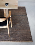 A wooden table with a black coffee cup, a wooden chair with a black seat, and the Armadillo&Co Drift Rug - Natural & Black on a light gray concrete floor showcase Fair Trade craftsmanship.