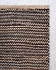 A close-up of the Drift Rug in Natural & Black by Armadillo&Co, featuring a wavy striped jute and wool weave in brown and black, placed on a light terrazzo floor.