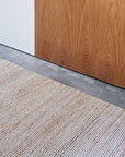 A close-up of the Armadillo&Co Drift Rug in Natural & White, hand-woven and flat weave, displayed on a concrete floor beside a light brown wooden door and a white paneled wall.