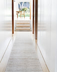 A bright, modern hallway with white walls features Armadillo&Co’s Drift Rug in Natural & White on wooden floors, leading up steps to a sunlit room with a wooden table and chair, overlooking green trees.