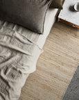 A neutral-toned bed with wrinkled linen sheets and two pillows sits beside the Armadillo&Co Drift Rug in Natural & White and a white stone nightstand holding a small dish, as sunlight creates soft shadows across the room.